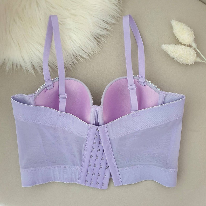 May include: A lavender-colored bustier-style bra with adjustable straps and a hook-and-eye closure. The bra features a mesh overlay and is adorned with small, pearl-like embellishments along the top edge of the cups.