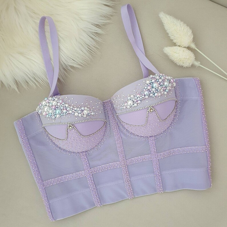 May include: A lavender corset top with delicate straps and intricate embellishments. The bust area is adorned with pearls and rhinestones, adding a touch of glamour. The corset features a structured design with vertical and horizontal lines, creating a flattering silhouette.