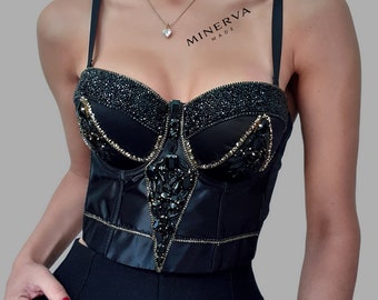 May include: Black satin bustier top with beaded embellishments. The top features a sweetheart neckline, a central zipper, and decorative gold stitching. The text "MINERVA MADE" is printed on the upper right side.