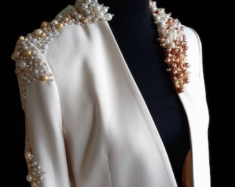 Puede incluir: Una chaqueta blanca con un adorno de perlas y cuentas doradas en el cuello y el hombro.