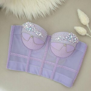 May include: A lavender corset top with intricate detailing. The bust area is adorned with pearls and rhinestones, and the boning is accented with rows of small, sparkling embellishments. The corset features a structured design.