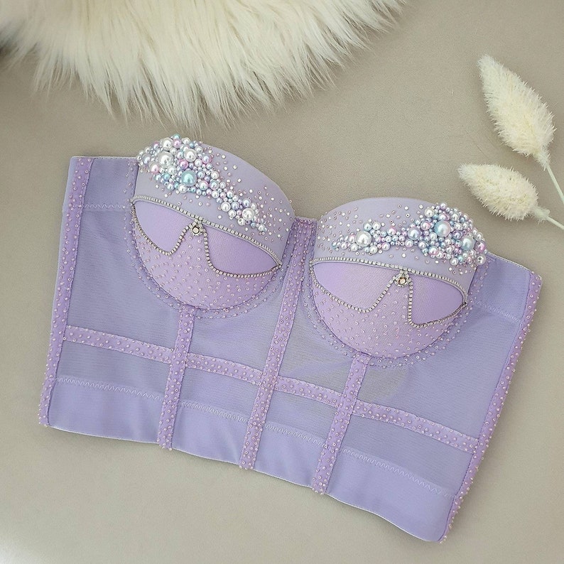 May include: A lavender corset top with intricate detailing. The bust area is adorned with pearls and rhinestones, and the boning is accented with rows of small, sparkling embellishments. The corset features a structured design.