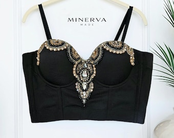May include: Black bustier top with intricate bead and jewel embellishments. The top features a sweetheart neckline and thin black straps. The brand name "MINERVA MADE" is visible at the top. The bustier is hanging on a white hanger.