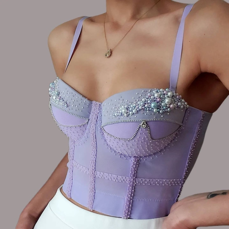 May include: A lavender corset top with delicate pearl and rhinestone embellishments. The bustier features a structured design with vertical and horizontal seams, and thin lavender straps. The top is a light purple color and has a subtle dotted texture.