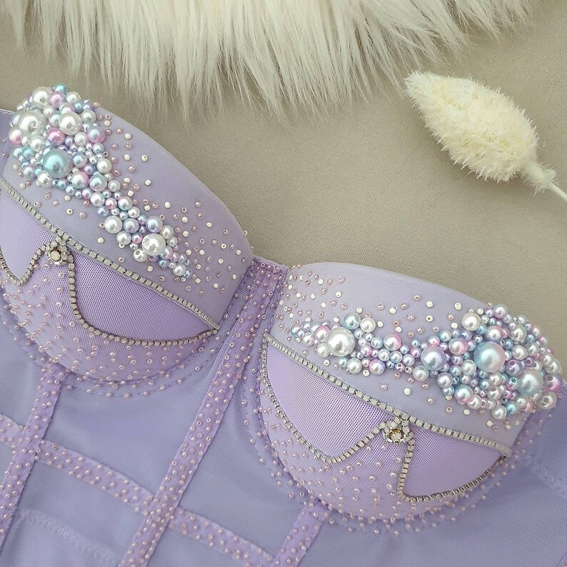 May include: A lavender corset top with pearl and rhinestone embellishments. The bust features clusters of pearls and iridescent beads. The corset has a structured design with vertical and horizontal lines, and a sheer overlay.
