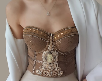 May include: A brown strapless corset with gold and white embellishments. The corset has a beaded design and a cameo-like pendant in the center.