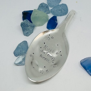 May include: A silver spoon with the text "Fell asleep here" and star designs. The spoon is surrounded by blue and green sea glass pieces on a white surface. A charming, decorative item.