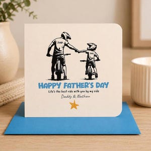 May include: A square greeting card with a cream background featuring a black illustration of two people on bicycles fist-bumping. Text reads: HAPPY FATHER'S DAY, Life's the best ride with you by my side, Daddy & Nathan, with a yellow star below.