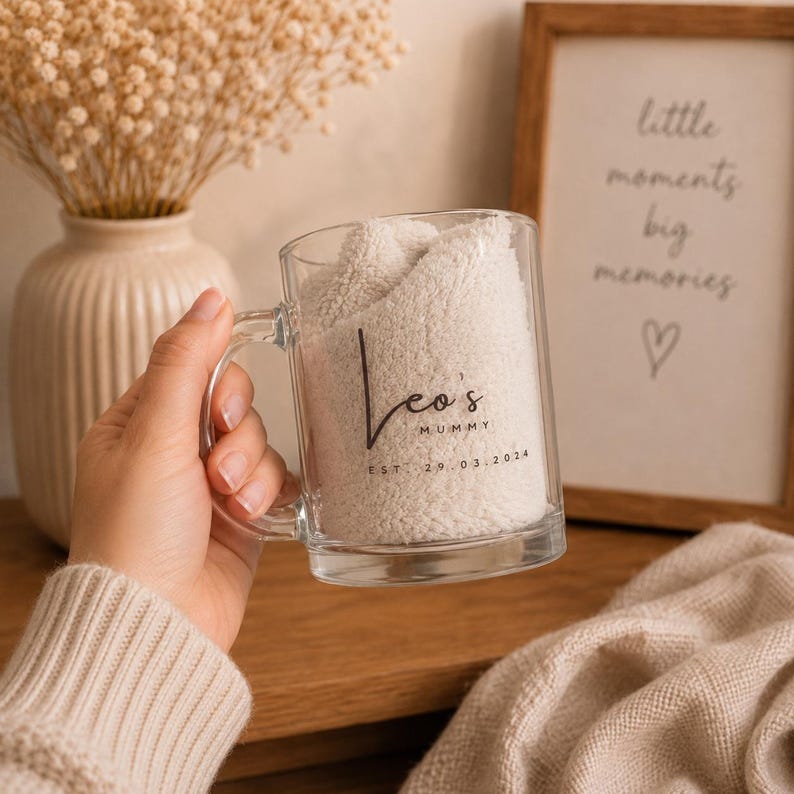 Personalised glass mug with custom mummy name and established date, held in a cosy neutral home setting with soft knitted blanket and dried flowers.