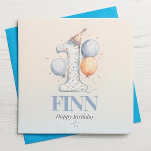 May include: A birthday card with a blue envelope. The card features a watercolor illustration of the number 1 with confetti, balloons, and a party hat. The name "FINN" and "Happy Birthday" are printed below the number.