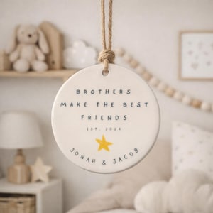 May include: A round white ceramic ornament hanging from a jute twine loop. The text reads BROTHERS MAKE THE BEST FRIENDS EST. 2024 with a yellow star graphic and the names JONAH & JACOB printed in dark blue lettering on the front surface.