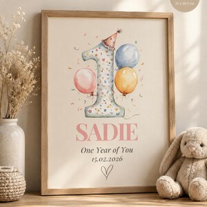 May include: A framed A4 print measuring 21 x 29.7 cm featuring a watercolor illustration of the number one wearing a party hat, surrounded by pink, blue, and yellow balloons. The text reads SADIE, One Year of You, 15.02.2026, and a heart.