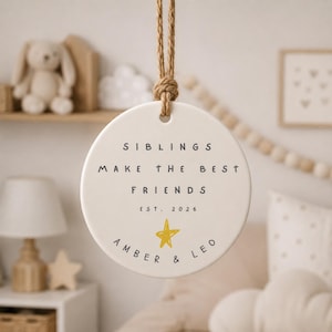 May include: A round white ceramic ornament hanging from a thick jute rope. The text reads SIBLINGS MAKE THE BEST FRIENDS EST. 2026 with a hand-drawn yellow star and the names AMBER & LEO printed in black lettering on the front surface.