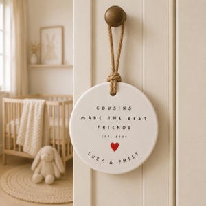 May include: A round white ceramic hanging plaque suspended by a thick jute rope from a brass knob. The plaque features black text that reads COUSINS MAKE THE BEST FRIENDS EST. 2026, followed by a small red heart and the names LUCY & EMILY.