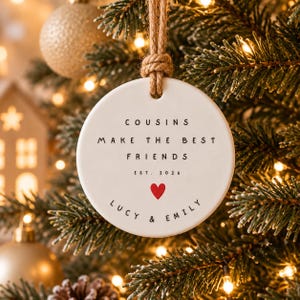 Puede incluir: Un adorno redondo de cerámica blanca que cuelga de un lazo de cuerda de yute. El texto dice "COUSINS MAKE THE BEST FRIENDS EST. 2026" con un pequeño icono de corazón rojo debajo y los nombres "LUCY & EMILY" impresos en negro en la base.