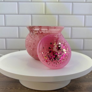 Puede incluir: Tarro de cristal rosa con purpurina y tapa. El tarro y la tapa son de color rosa con purpurina dorada incrustada en la resina.