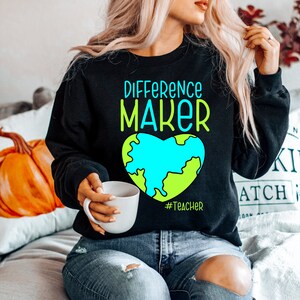 Puede incluir: Sudadera negra con el texto "Difference Maker" en letras blancas y azules. Debajo del texto hay un globo terráqueo en forma de corazón verde y azul. El texto "#Teacher" está debajo del globo.