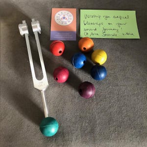 May include: A silver tuning fork with a handle and two prongs, labeled "OM 136.1".  The tuning fork is surrounded by seven colorful balls: red, orange, yellow, green, blue, purple, and teal.
