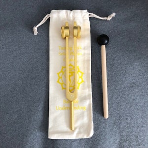 Puede incluir: Un diapasón dorado con un mazo de madera en una bolsa de tela blanca. El diapasón tiene una etiqueta amarilla con el texto "Tuning Fork Solar Plexus Chakra Balance Understanding".