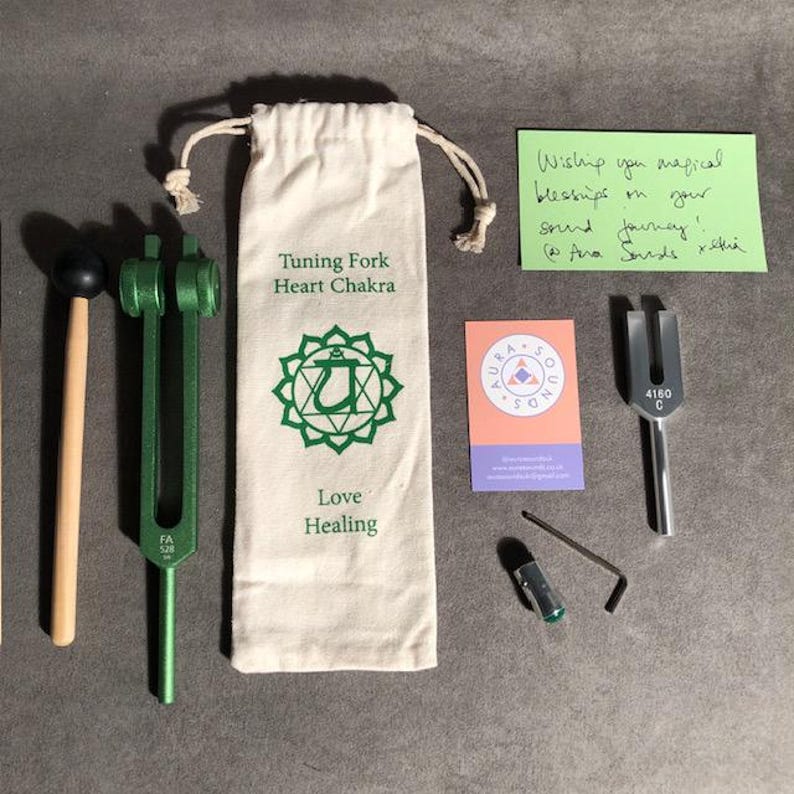 K&ouml;nnte beinhalten: Eine gr&uuml;ne Stimmgabel f&uuml;r das Herzchakra, mit einem Holzhammer, einem kleinen Beutel und einer Karte mit der Aufschrift "Aura Sounds". Die Stimmgabel ist mit "4160 C" und "FA 528" beschriftet. Der Beutel hat ein gr&uuml;nes Herzchakra-Symbol und die Worte "Tuning Fork Heart Chakra", "Love Healing".