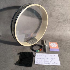 May include: A wooden frame drum with a clear plastic head, a black beaded bracelet with rainbow colored beads, a tuning fork, and a card with the text "Wishing you magical blessings on your sound journey!  - Anna Sounds India".