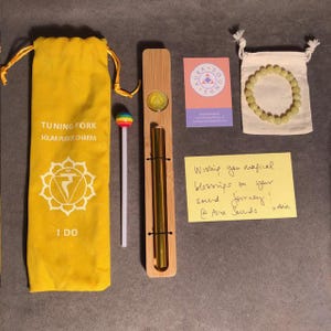 Puede incluir: Una bolsa de terciopelo amarilla con las palabras "Tuning Fork Solar Plexus Chakra" y el símbolo del chakra del plexo solar. También se ve un palo de piruleta arcoíris, un diapasón dorado en un soporte de madera, una pequeña bolsa con un brazalete de piedra verde y una tarjeta con las palabras "Aura Sounds". También se incluye una nota adhesiva amarilla con el texto "Wishing you magical blessings on your sound journey! @Ava Sounds xox".