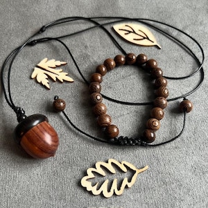 May include: A collection of wooden jewelry and decorative elements. Includes a necklace with an acorn pendant, a beaded bracelet, and several laser-cut wooden leaf shapes. The items are brown and black, set against a gray background.