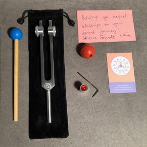 May include: A collection of sound healing tools, including a silver tuning fork with a rose emblem, a blue mallet, a red sphere, a small red crystal, and a business card. A handwritten note reads "Wiship you refical blessings on your sound Jouney! @Aura Sounds tana."