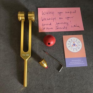 May include: A gold tuning fork with a red mallet and a small wrench. The tuning fork is labeled "194.18HZ". A business card for Aura Sounds is also included.