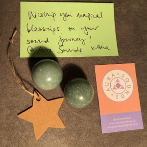 May include: Two green, round, textured sound healing singing bowls with a brown star-shaped tag and a business card with the text "Aura Sounds" in a purple triangle on a peach background.