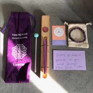 Puede incluir: Una bolsa de terciopelo morado con las palabras "Tuning Fork Crown Chakra" e "I Know" impresas en ella. Un palo blanco con una bola azul y blanca a rayas en la parte superior. Un soporte de madera con un diapasón morado en su interior. Una pequeña bolsa blanca con un brazalete de amatista morado dentro. Una tarjeta rosa con las palabras "Aura Sound" impresas en ella. Una nota escrita a mano que dice "Wishing you magical blessings on your sound journey! @Aura Sounds x Aura"