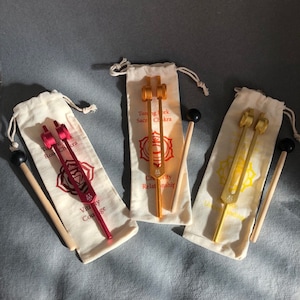 May include: Three tuning forks in white drawstring bags with wooden mallets. The tuning forks are red, orange, and gold. The bags have text that reads "Tuning Fork Sacral Chakra Creativity Relationship", "Tuning Fork Root Chakra Vitality Courage", and "Tuning Fork Solar Plexus Grounding".
