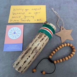 May include: A wooden incense holder with a green, red, and white band, a wooden bead bracelet, and a star-shaped tag. A yellow note reads "Wishing you magical blessings on your sand journey! @Aura Sounds x sna". A pink and white business card is also present.