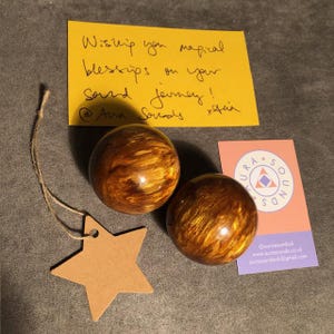 Puede incluir: Dos bolas de sonido marmoleadas de color marrón y dorado con una etiqueta en forma de estrella marrón y una tarjeta de visita con el texto "Aura Sounds" y la información de contacto.