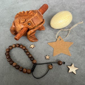 Pode incluir: Uma coleção de itens de madeira, incluindo um instrumento em forma de sapo, uma pulseira com contas marrons escuras, um ovo de madeira de cor clara, uma etiqueta em forma de estrela e pequenas estrelas de madeira. O instrumento de sapo tem um pau para fazer um som.