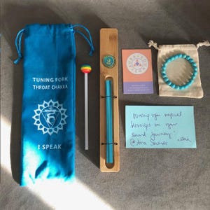 Puede incluir: Una bolsa de terciopelo azul con las palabras "Tuning Fork Throat Chakra" y "I Speak" impresas en ella. Una caja de madera con un diapasón azul dentro. Un palo de piruleta de colores arcoíris. Una pulsera turquesa en una pequeña bolsa blanca. Una tarjeta con las palabras "Aura Sounds" impresas en ella. Una nota adhesiva azul con las palabras "Wiring you magical blessings on your sound journey! x Ava Sounds" escritas en ella.