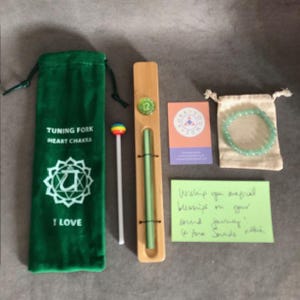 Puede incluir: Una bolsa de terciopelo verde con las palabras "Tuning Fork Heart Chakra" e "I Love" impresas en ella. Un palo blanco con una bola de colores arcoíris en la parte superior. Una caja de madera con un cristal verde en su interior. Una pequeña bolsa con un brazalete de cuentas verdes. Una tarjeta con las palabras "Aura Sound Journey" impresas en ella. Una nota escrita a mano que dice "Wishing you magical blessings on your sound journey.  Love, Sandra & Allen."