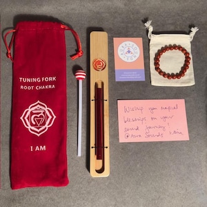 Puede incluir: Una bolsa de terciopelo rojo con las palabras "Tuning Fork Root Chakra" e "I AM" impresas en ella. Un soporte de madera con un diapasón rojo en su interior. Un mazo de diapasón a rayas blancas y rojas. Un brazalete rojo en una bolsa de cordón blanco. Una nota rosa con el texto "Wish you magical blessings on your sound journey! @Aura Sounds kana".