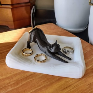 May include: A black cat-shaped ring holder with a white tray, holding several gold and silver rings. The cat is in a stretching pose, with its tail arched. The tray sits on a wooden surface.