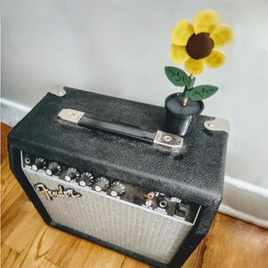 Puede incluir: Un amplificador de guitarra Fender negro con una decoraci&oacute;n de girasol en la parte superior. El amplificador tiene una placa frontal plateada con botones y la palabra "Fender" impresa en ella. El girasol es amarillo con un centro marr&oacute;n y hojas verdes.