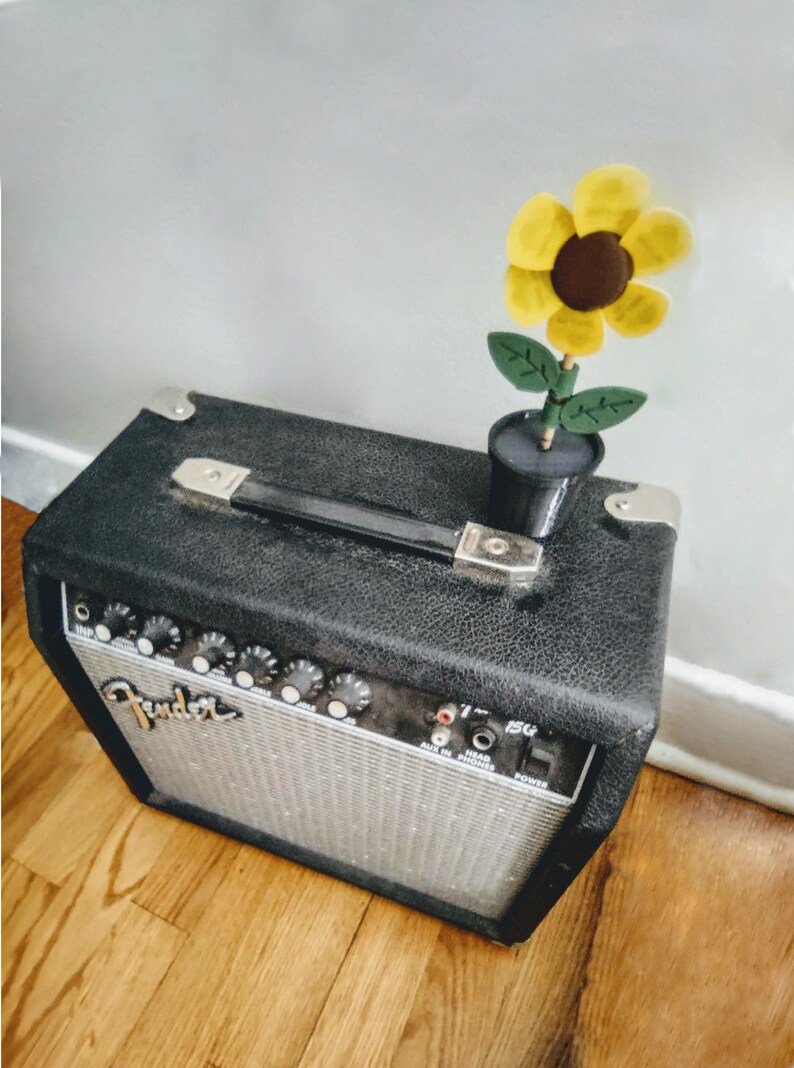 Puede incluir: Un amplificador de guitarra Fender negro con una decoraci&oacute;n de girasol en la parte superior. El amplificador tiene una placa frontal plateada con botones y la palabra "Fender" impresa en ella. El girasol es amarillo con un centro marr&oacute;n y hojas verdes.