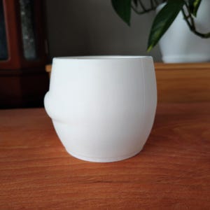 May include: A white ceramic plant pot with a rounded shape and a small, curved detail on one side. The pot is on a wooden surface, with a blurred background including a cabinet and a houseplant in a white pot.