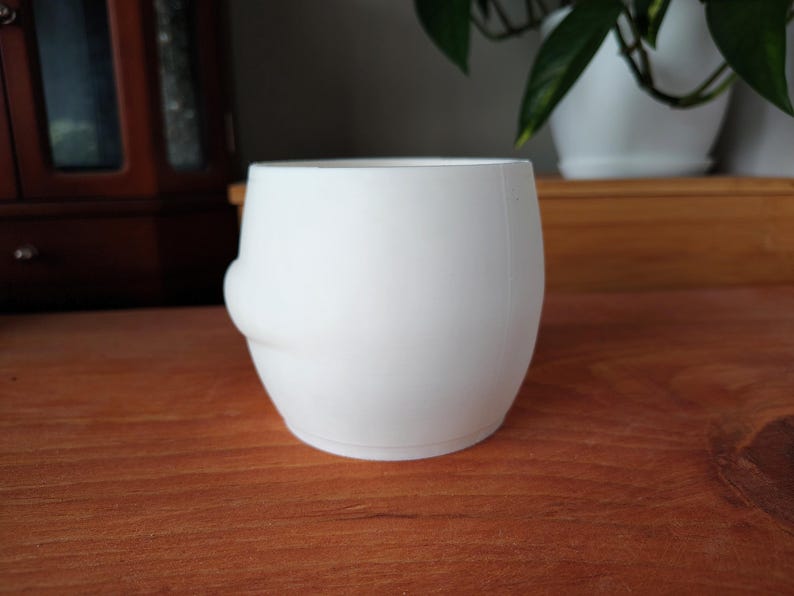 May include: A white ceramic plant pot with a rounded shape and a small, curved detail on one side. The pot is on a wooden surface, with a blurred background including a cabinet and a houseplant in a white pot.