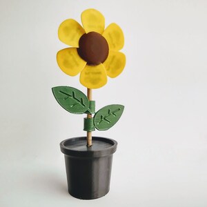 Puede incluir: Un girasol amarillo con un centro marr&oacute;n est&aacute; unido a un palo de madera y colocado en una maceta negra. El girasol tiene hojas verdes.