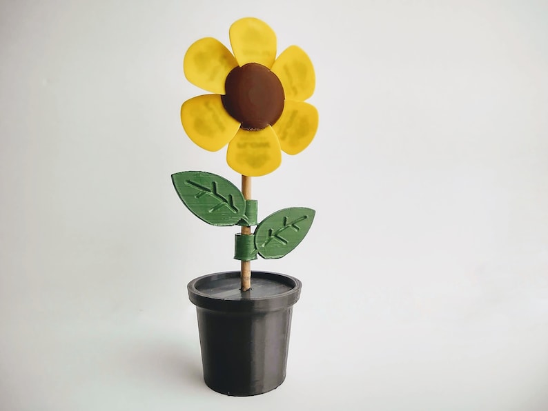 Puede incluir: Un girasol amarillo con un centro marr&oacute;n est&aacute; unido a un palo de madera y colocado en una maceta negra. El girasol tiene hojas verdes.