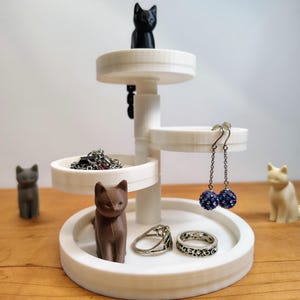 May include: A white, three-tiered jewelry organizer with a cat figurine on top and several cat figurines around the base. The organizer holds rings, earrings, and a necklace. The earrings have blue beaded accents.