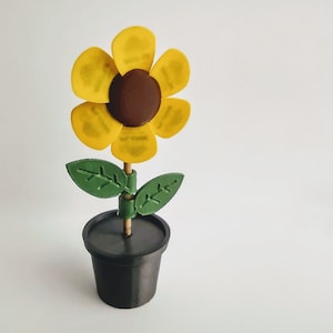 Puede incluir: Un girasol amarillo con un centro marr&oacute;n est&aacute; en una maceta negra con hojas verdes. El girasol est&aacute; hecho de papel y es un proyecto artesanal.