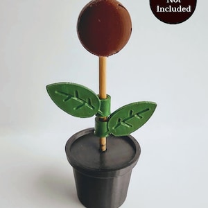 Puede incluir: Una flor marr&oacute;n impresa en 3D con hojas verdes, sentada en una maceta negra. La flor est&aacute; en un palo de madera. "Picks Not Included" est&aacute; escrito en un c&iacute;rculo negro en la esquina superior derecha de la imagen.