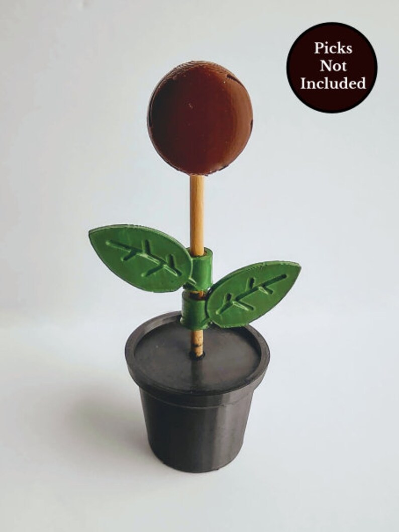 Puede incluir: Una flor marr&oacute;n impresa en 3D con hojas verdes, sentada en una maceta negra. La flor est&aacute; en un palo de madera. "Picks Not Included" est&aacute; escrito en un c&iacute;rculo negro en la esquina superior derecha de la imagen.