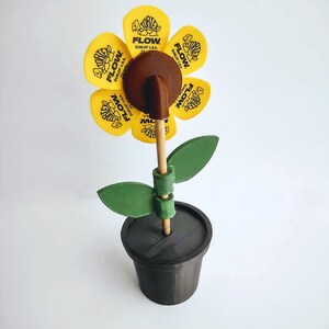 Puede incluir: Una flor amarilla hecha de p&uacute;as de guitarra con el texto "FLOW" y "DUNLOP U.S.A." en cada p&uacute;a. La flor est&aacute; en un palo marr&oacute;n y en una maceta negra con hojas verdes.